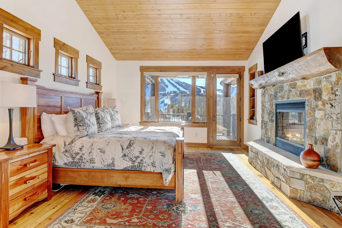A serene primary suite featuring a king-sized bed with inviting patterned bedding, a stone fireplace, and expansive windows that frame breathtaking mountain views. The soft natural light and cozy textures create a peaceful atmosphere, perfect for rest and romance.