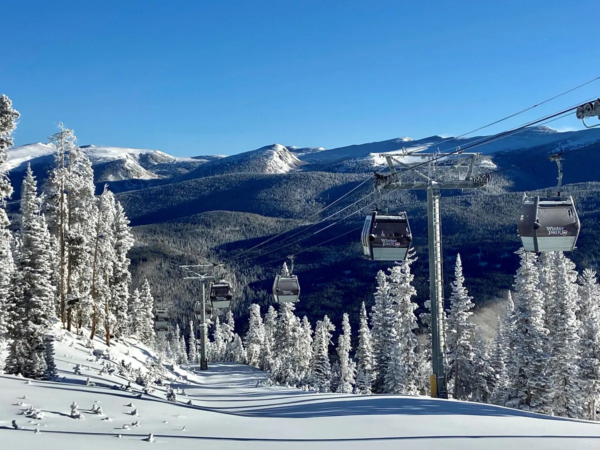 A scenic winter landscape featuring ski gondolas gliding above snow-covered trees and distant mountain peaks. The image captures the exhilarating atmosphere of Winter Park Resort, with clear blue skies and pristine snow evoking the thrill of winter sports.