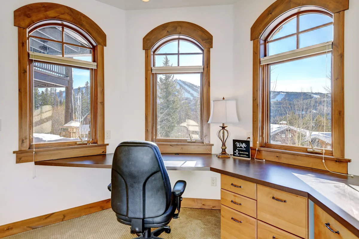 A serene office area with a wooden desk, comfortable office chair, and large windows framing stunning mountain views. Ample natural light and a warm, productive atmosphere make this space ideal for remote work or quiet relaxation.