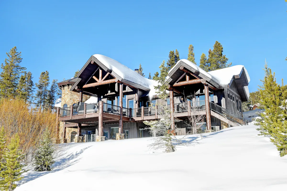 A view of the home's wooden exterior, featuring expansive windows overlooking a snowy landscape and forested surroundings. The deck is a focal point, inviting thoughts of both winter sports and tranquil moments, with the warmth inside suggested by the visible fireplace glow.
