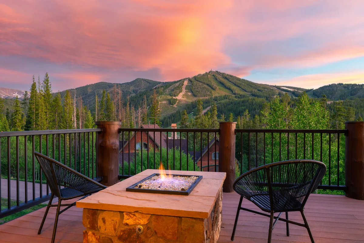 An inviting outdoor fire pit setup on a spacious deck, surrounded by comfortable chairs and offering a panoramic mountain view. The sky above is dotted with clouds, enhancing the sense of tranquility and open space. The scene conveys a cozy yet invigorating connection with nature, perfect for relaxing or taking breaks from work.