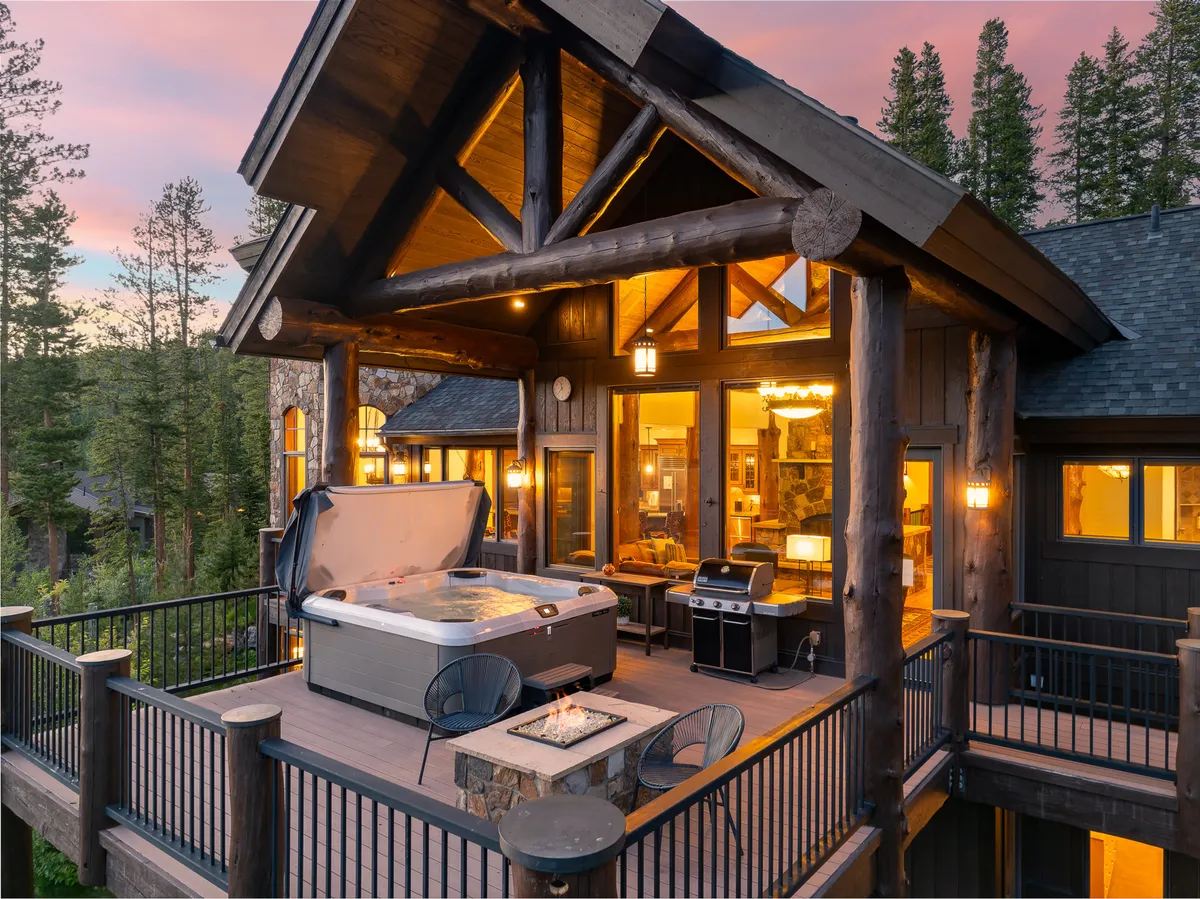 A tranquil, luxurious exterior patio scene featuring a private hot tub, outdoor fire pit, dining area, and wooden architectural beams. The setting sun casts a golden glow, inviting guests to relax after a day of adventure.