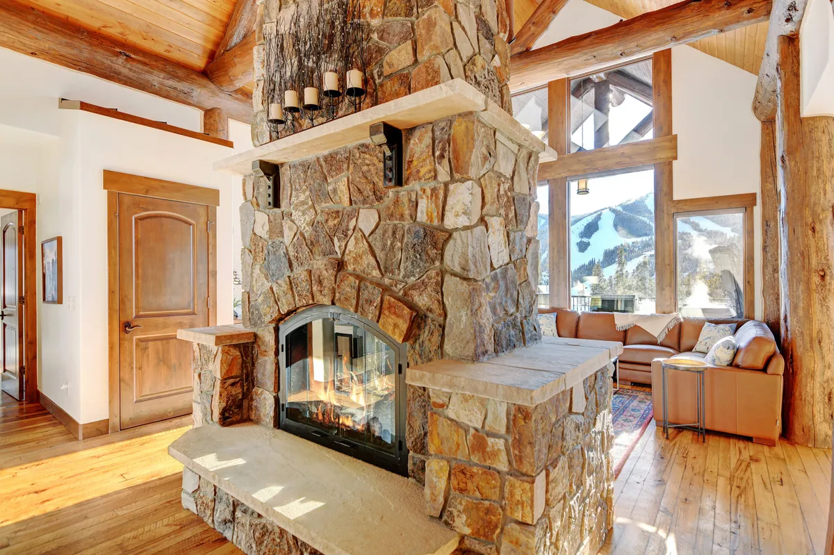 A cozy seating area in the living room featuring a dramatic stone fireplace as the focal point, surrounded by large windows that offer sweeping views of the surrounding mountains. Rustic wooden beams accentuate the vaulted ceilings, while plush seating invites relaxation for groups or families.