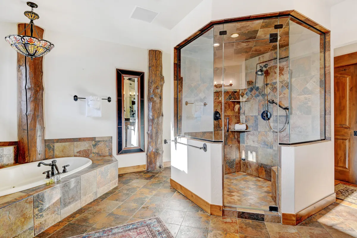A luxurious bathroom highlighted by a freestanding soaking tub under a decorative chandelier, a glass shower enclosure, natural wood beams, and a mirrored wall. The room exudes spa-like tranquility, inviting relaxation and indulgence.