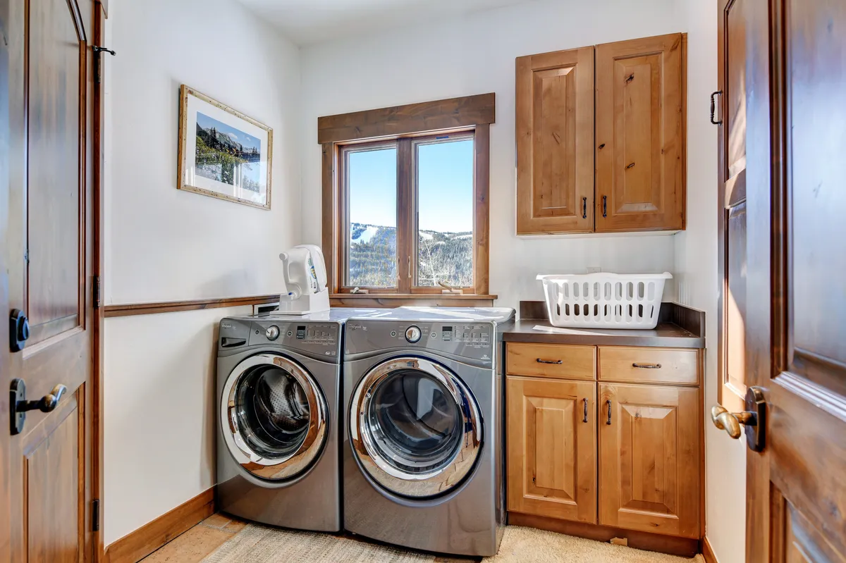 A bright laundry area with modern washing machines, large windows revealing a mountain view, and warm wood cabinetry. The practical yet inviting space suggests convenience for extended stays and daily routines, all while keeping a visual connection to the outdoors.