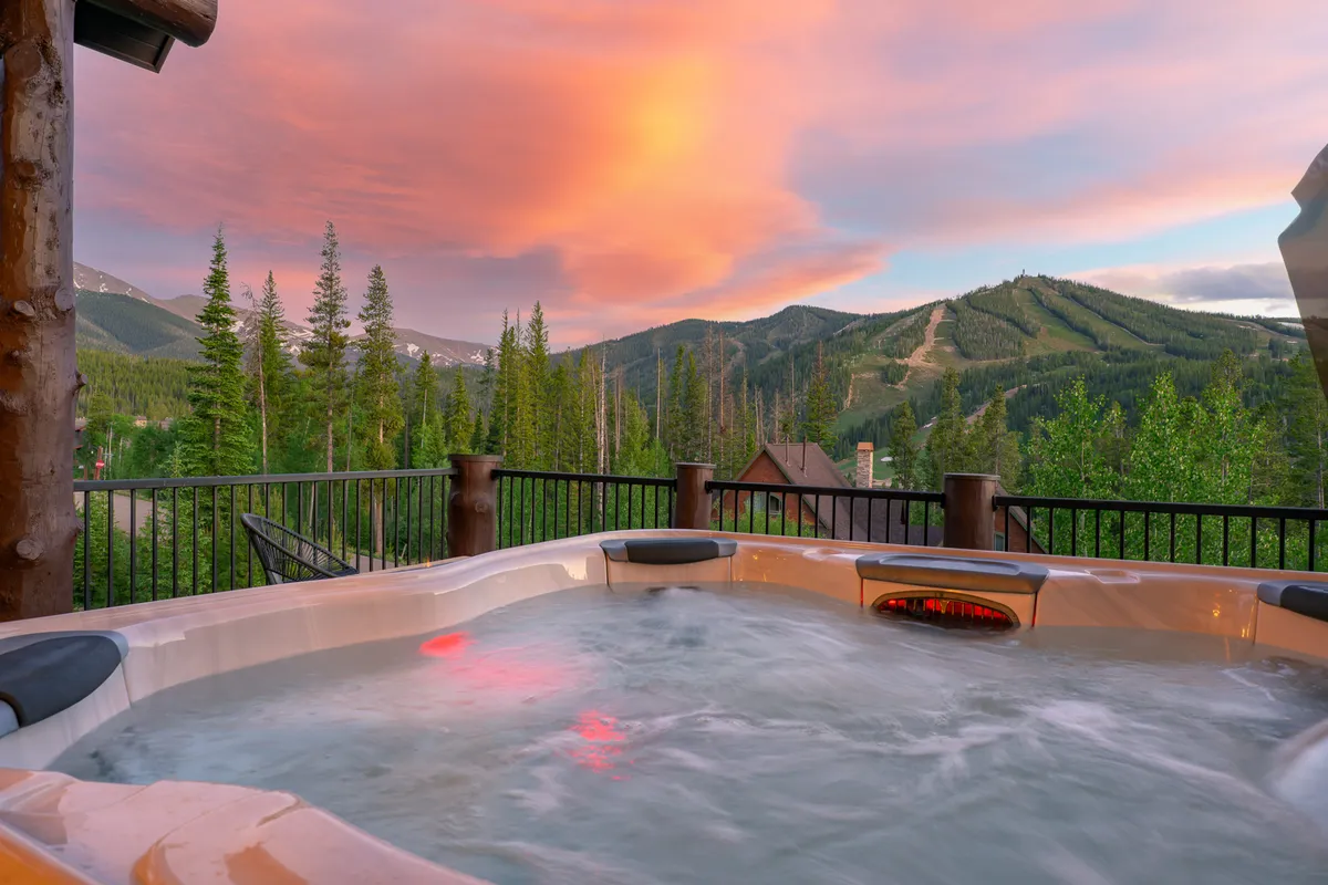 This image captures the outdoor hot tub with a sweeping view of the mountain range under a brilliant sunset sky. Guests are seen relaxing in the warmth of the water, soaking in the tranquil atmosphere and panoramic scenery.