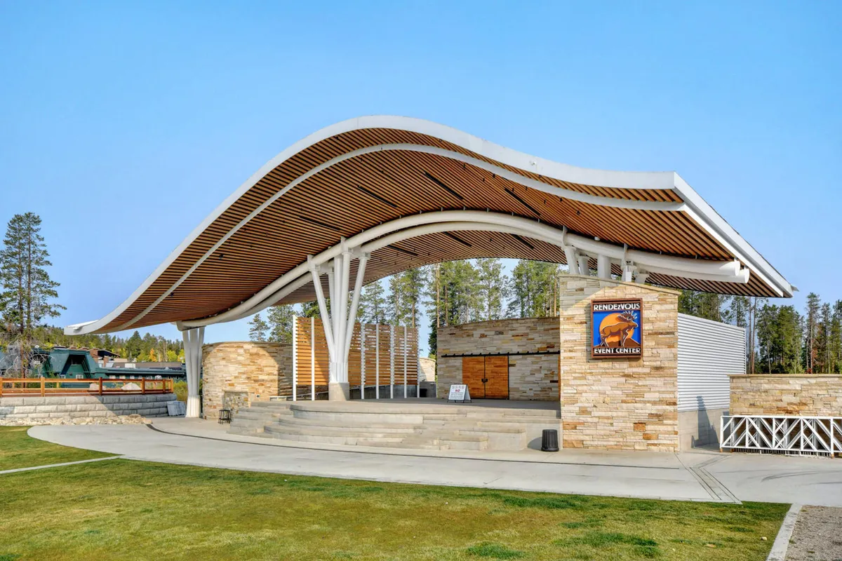 The exterior shot features a distinctive wave-shaped roof, stone facade, and signage indicating a nearby event center, with open skies above. This scene highlights the resort-like amenities and opportunities for exclusive adventures in the area.