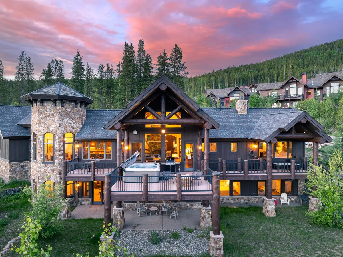 A spacious outdoor deck showcasing a large hot tub, stone exterior, and floor-to-ceiling windows. Surrounded by trees, the deck offers ample space for outdoor dining, family gatherings, and relaxation while overlooking the beautiful natural setting.