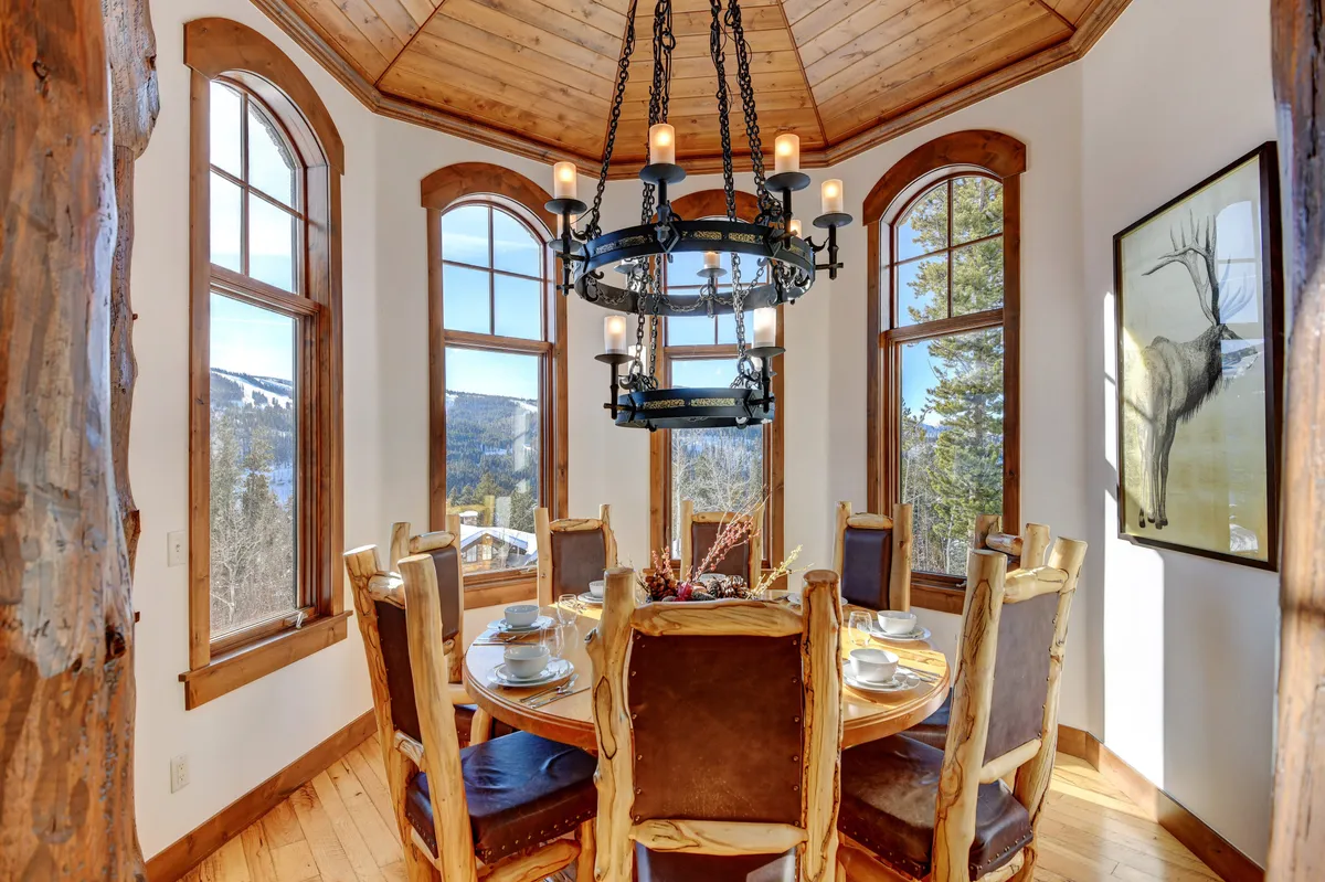 A spacious, warmly lit dining area featuring a long wooden table surrounded by custom log chairs, set beneath an elegant chandelier. Large windows wrap the space, offering 360-degree views of the surrounding mountains, creating a celebratory setting for shared meals.