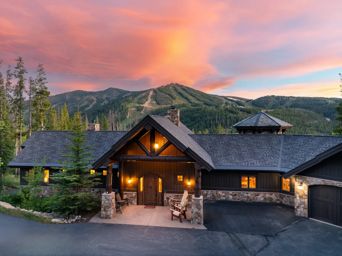 A welcoming view of the home's front entrance, featuring a sturdy wooden door framed by stone pillars. Warm lights glow from inside, with expansive mountain vistas in the background, inviting guests to explore or gather outside as dusk falls.