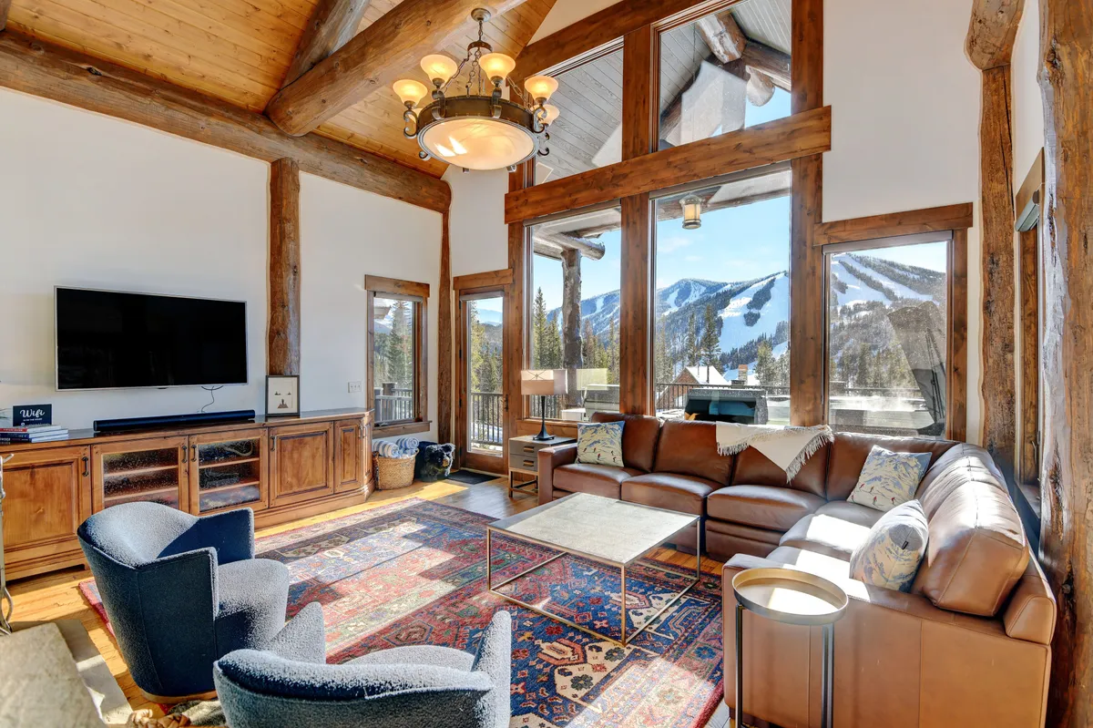 A welcoming living room with a leather sofa, colorful area rug, rustic wooden beams overhead, and expansive windows that frame stunning mountain vistas. The arrangement invites conversation, relaxation, and enjoying cozy moments together.