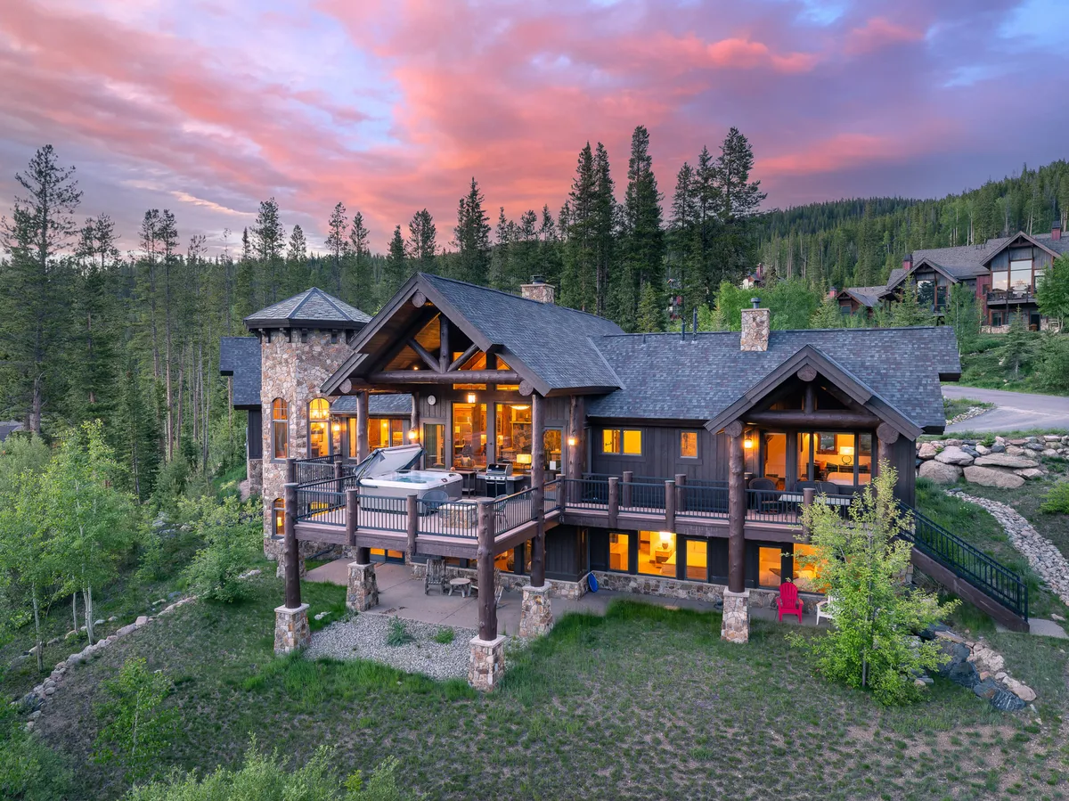 The image showcases the villa's striking stone and timber architecture, a spacious balcony with comfortable seating, and expansive windows reflecting the vibrant hues of a mountain sunset. The serene outdoor space invites guests to gather for sunset views, enjoy morning coffee, or unwind in the private hot tub, all set against the backdrop of Winter Park's natural beauty.