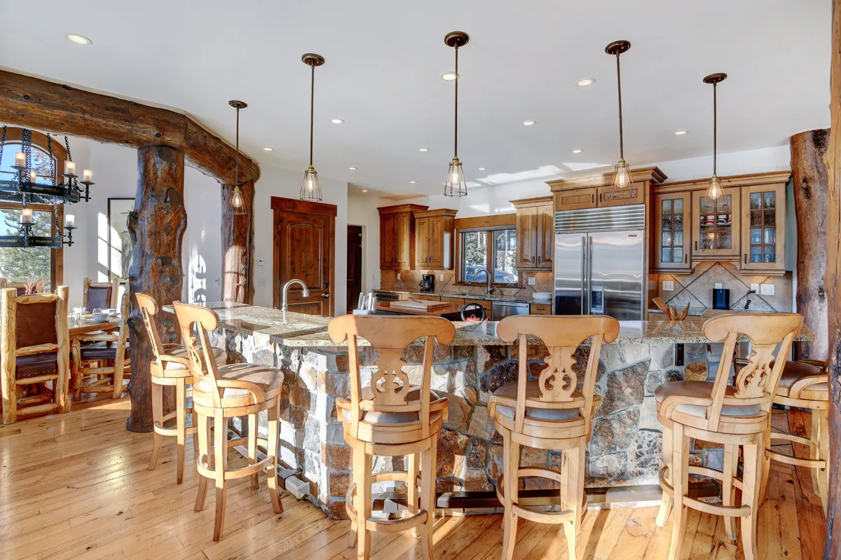 A warmly lit kitchen featuring two spacious islands topped with elegant stone countertops, surrounded by bar stools and wooden beams overhead. The space buzzes with the potential for entertaining, family gatherings, and group meal preparation, with ample seating at the islands and a cozy, inviting atmosphere.